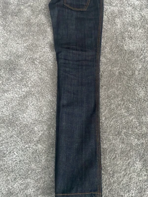 Nudie jeans dry denim - Säljer ett par mörkblå jeans från Nudie Jeans med klassisk orange söm och vågig detalj på bakfickan. Perfekta för dig som gillar stilrena och tidlösa jeans. De är i dry denim så de får snygga fades efter användning. Storlek w29L32.          Kan vara bra att gå ner i storlek då jeansen töjs ut då de används mycket och inte tvättas.