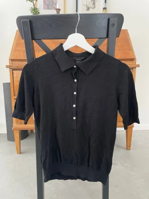 Svart polotopp från Lindex - Klassisk svart polotopp från Lindex med korta ärmar och knappar framtill. Toppen har en mjuk krage och ribbad nederkant, vilket ger en stilren och enkel look. Perfekt att matcha med jeans eller kjol för en clean outfit.
