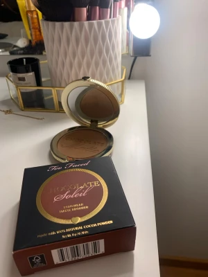 Too Faced Chocolate Soleil Bronzer - Säljer en Too Faced Chocolate Soleil Longwear Matte Bronzer i en snygg guldfärgad rund dosa med spegel. Bronzern har en matt, varm brun ton och är gjord med naturligt kakaopulver för en lyxig känsla och doft. Perfekt för att skapa en solkysst look.