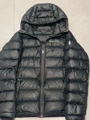 Svart dunjacka från Peak Performance - Snygg svart dunjacka från Peak Performance med huva och dragkedja framtill. Jackan har quiltad design, diskret logga på bröstet och en ficka på ärmen. Perfekt för kalla vinterdagar och riktigt skön att bära. Dragkedjan är inte så bra nästan att den behövs byta