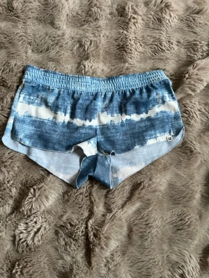 Blå tie-dye shorts från Roxy - Säljer ett par blåvita tie-dye shorts från Roxy med klassisk femficksdesign och röda detaljer på bakfickan. Shortsen har låg midja och är perfekta för sommardagar. Sköna och snygga att matcha med en hoodie eller t-shirt.