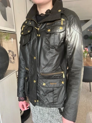Svart vaxad klassisk jacka från Barbour - Snygg svart vaxad och fodrad jacka från Barbour med guldiga detaljer och flera fickor med dragkedjor och knappar. Klassisk krage och figurnära passform. Knappt använd pga liten i storleken. Är strl 38 men skulle mer säga att det passar strl 36. 