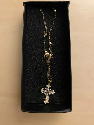 Guldigt halsband 50cm med korshänge - Snyggt halsband i guldfärgad metall med ett korsformat hänge dekorerat med gnistrande stenar. Kedjan har en tvinnad design som ger extra bling och hänget är detaljrikt med vita stenar som fångar ljuset. Perfekt accessoar för dig som gillar statement-smycken.