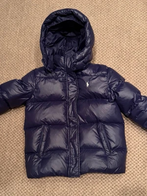 Mörkblå vinterjacka från Polo Ralph Lauren - Snygg mörkblå vinterjacka från Polo Ralph Lauren med huva och dragkedja. Jackan har två snedställda fickor framtill och den klassiska Polo-loggan broderad på bröstet. Perfekt för kalla vinterdagar. Jackan är sparsamt använd en säsong och i mycket bra skick. 