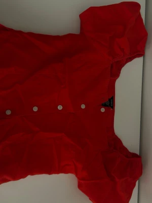 Röd blus med puffärm från Lindex - Snygg röd blus från Lindex med puffiga korta ärmar och vita knappar framtill. Tillverkad i ett lätt tyg som känns skönt mot huden. Perfekt för dig som vill ha en färgstark och trendig look❤️