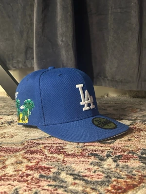 Blå LA Dodgers keps från New Era - Säljer en blå snapback-keps från New Era med LA Dodgers-logga broderad framtill i vitt och palmträd med texten 'Greetings from LA' på sidan. Klassisk rak skärm och broderade ventilationshål. Perfekt för dig som vill ha en sportig och trendig look.