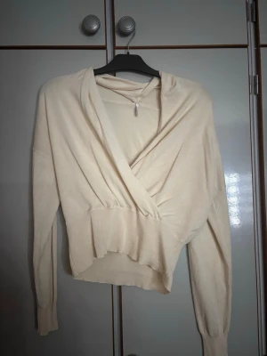 Beige omlottopp med lång ärm - Snygg beige omlottopp med lång ärm och bred mudd i midjan. Tröjan har en djup omlottringning och är tillverkad i ett mjukt, stretchigt material. Perfekt för en trendig och avslappnad look.