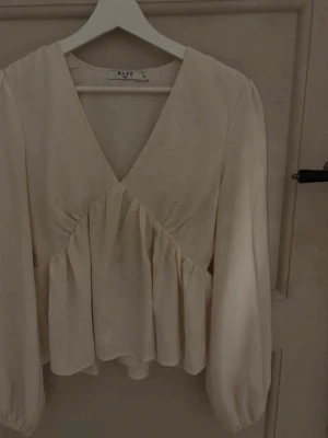 Krämvit blus från NA-KD med v-ringning - Supersnygg krämvit blus från NA-KD med djup v-ringning och volang nedtill. Blusen har långa ärmar med lätt puff. Storlek 32. Inte använd så många gånger 💗