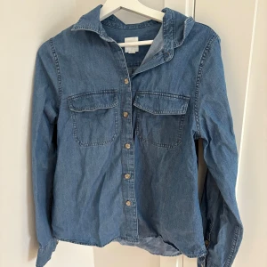 Blå jeansskjorta med bröstfickor - Klassisk blå jeansskjorta i bomull med två bröstfickor och knappar framtill. Skjortan har lång ärm och en avslappnad passform, perfekt att styla lager på lager eller bära som den är. Tidlös och enkel att matcha till olika outfits.