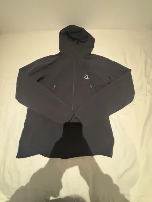 Svart vindjacka från Haglöfs med huva - Snygg svart vindjacka från Haglöfs med hel dragkedja, två sidofickor med dragkedja och justerbar huva. Jackan har minimalistisk design och Haglöfs-logga på bröstet. Perfekt för blåsiga dagar och outdoor-äventyr.