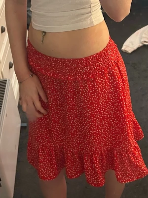 Röd kort/lång kjol  - Superfin röd kortkjol med vitta prickar och volangkant. Kjolen har resår i midjan för en bekväm passform och är perfekt för varma dagar. Lätt och luftig modell som ger en söt och trendig look.