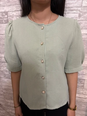 Ljusgrön blus med puffärm och pärlknappar - Snygg ljusgrön blus med puffiga korta ärmar och rund halsringning. Blusen har dekorativa pärlknappar framtill och är gjord i ett strukturerat tyg som ger en trendig look. Perfekt för dig som vill ha en söt och stilren topp till vardagsoutfiten.
