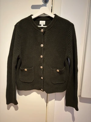 Mörkgrön stickad kofta med fickor - Säljer en mörkgrön stickad kofta med rund halsringning och träknappar framtill. Koftan har två framfickor med knappdetaljer och ribbade muddar vid ärmslut och nederkant. 