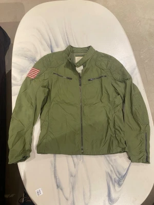 Denim and supply ralph lauren field jacket - Snygg grön vindjacka med ståkrage och dragkedja framtill. Två bröstfickor med dragkedja och quiltade detaljer på axlarna. Amerikansk flagga som patch på höger ärm ger jackan en cool militärinspirerad look. Perfekt för dig som gillar streetwear.