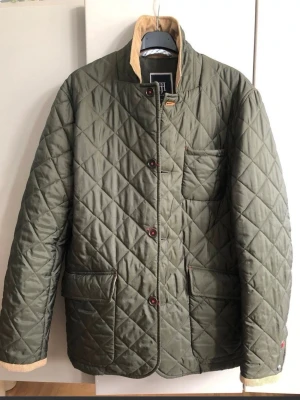 Olivgrön quiltad jacka Hansen & Jacob S/M  - Snygg olivgrön quiltad jacka från Hansen & Jacob med beige manchesterkrage och detaljer. Jackan har tryckknappar, tre fickor och ett klassiskt rutmönster. Perfekt för dig som gillar stilren och tidlös design. Ny 