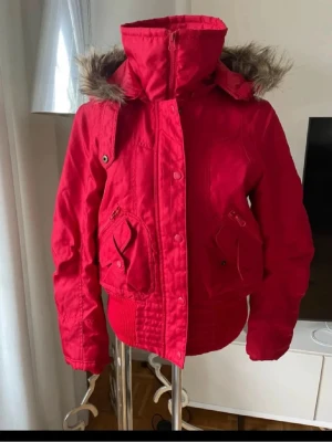 Röd bomberjacka med fuskpäls - Snygg röd bomberjacka med hög krage och avtagbar huva med fuskpäls. Jackan har två stora fickor framtill med lock och dragkedja samt ribbade muddar i midja och ärmslut. Perfekt för kyliga dagar och ger en cool streetstil.