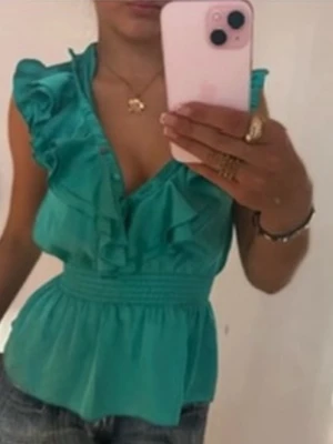 Turkos volangblus med v-ringning - Snygg turkos blus (vintage) med v-ringning och volangdetaljer framtill. Blusen har bred smock i midjan som ger en feminin siluett och är ärmlös, perfekt för varma dagar. Passar till både jeans och kjol för en trendig look. 