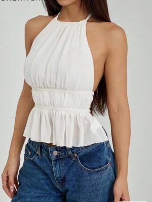 Vit halterneck topp från SHEIN - Trendig vit halterneck topp från SHEIN med smockad midja och volang nedtill. Toppen har rynkade detaljer och är ärmlös med öppen rygg, vilket ger en luftig och somrig känsla. Tillverkad i ett lätt och mjukt tyg som sitter snyggt på kroppen.