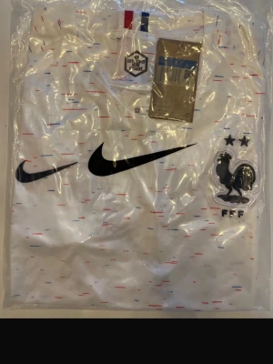 Frankrike 2018 Nike fotbollströja vit - Helt ny! Vit fotbollströja från Nike med Frankrikes landslagslogga och två stjärnor på bröstet. Tröjan har svarta Nike-loggor och små röda och blå detaljer över hela tyget. Perfekt för dig som älskar fotboll och vill stötta Les Bleus.