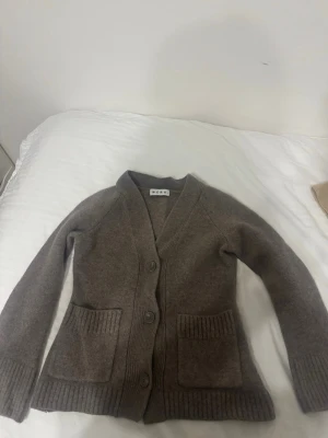 Brun kofta från Wera med fickor - Mysig brun kofta från Wera med djup V-ringning, tre knappar framtill och två stora fickor. Koftan har ribbstickade muddar och är tillverkad i ett mjukt, stickat material. Perfekt för lager-på-lager och chill dagar. Säljer pga krympning i tvätten!