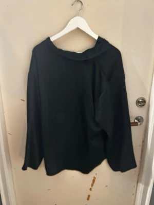 Svart blus med volangdetaljer - En svart blus med lång ärm och volangdetaljer vid axlar, halsringning och ärmslut. Blusen har en lös passform och hög hals. Perfekt för dig som gillar en stilren men ändå trendig look.