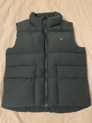 Svart dunväst från Gant - Stilren marinblå dunväst från Gant med hög krage och dragkedja framtill. Västen har diskret logga på bröstet och breda, quiltade paneler som ger en modern look. Perfekt för lager-på-lager och håller dig varm under kyliga dagar.