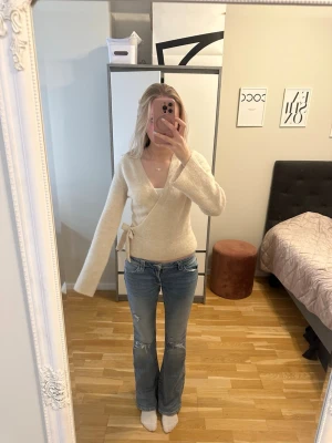 Beige omlottkofta med knytband  - Mysig beige omlottkofta med knytband i sidan och djup v-ringning. Koftan har långa ärmar och är stickad i ett mjukt material som ger en avslappnad look. Endast använd ett fåtal gånger.