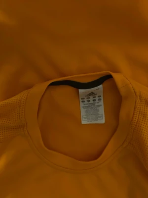 Orange Adidas sporttröja - Säljer en orange sporttröja från Adidas med svarta detaljer och klassiska ränder på axlarna. Tröjan är tillverkad i ett lätt och ventilerande syntetmaterial, perfekt för träning och aktivitet. Rund halsringning och korta ärmar.