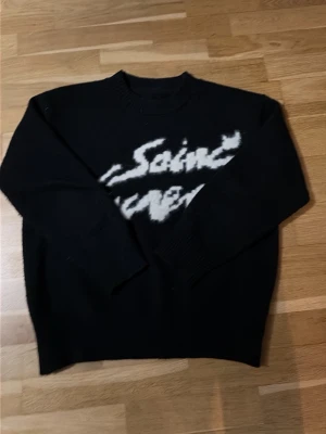 Svart stickad tröja Saint Laurent - Cool svart stickad tröja från Saint Laurent med vit fuzzy logga framtill. Tröjan har rund halsringning och långa ärmar. Perfekt för dig som gillar streetstyle och vill ha något trendigt och bekvämt i garderoben.
