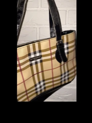 Burberry London väska! - Snygg handväska från Burberry med klassiskt rutigt mönster i beige, svart, vitt och rött. Väskan har svarta handtag i glansigt material och en rektangulär form. Perfekt för dig som gillar ikoniska designer och stilrena accessoarer.