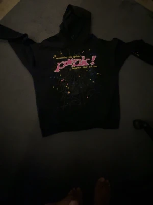 Svart sp5der hoodie ”P*NK” - Svart hoodie med huva och färgglatt tryck på bröstet där det står 'pink!' i rosa med vita prickar, samt texten 'nevermind the spider, heres the slime'. Motiv med stjärnor och spindelnät. Perfekt för dig som gillar unika och lekfulla plagg.