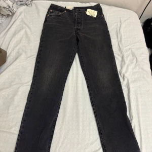 Svarta Levi's 501 jeans med knappar - Klassiska svarta Levi's 501 jeans med raka ben och hög midja. Jeansen har knappgylf och fem fickor, samt kopparfärgade nitar. Tillverkade i kraftig denim som ger en tidlös look. Perfekta för dig som gillar vintage vibe och ikonisk stil.
