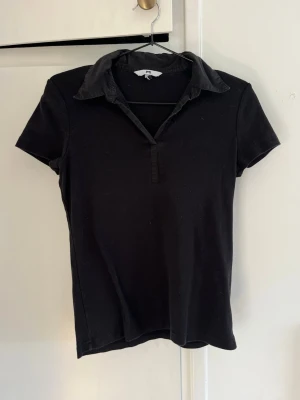 Svart kortärmad polotopp  - Klassisk svart polotopp  med korta ärmar och krage. Toppen har en enkel design med liten v-ringning och mjukt bomullsmaterial som gör den bekväm och lätt att matcha till olika outfits.