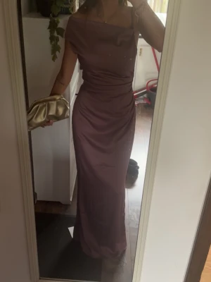 Lång lila axelbandslös klänning - Säljer en elegant långklänning i lila med axelbandslös design och draperad överdel. Klänningen har en figurnära passform och är perfekt för speciella tillfällen. Materialet har en lätt glansig finish som ger en lyxig känsla. Den är från Adoore och är aldrig använd. 