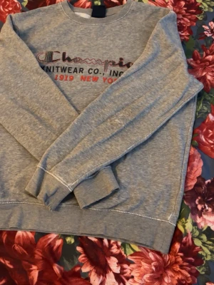 Grå stickad sweatshirt från Champion - Grå stickad sweatshirt från Champion med broderad logga och texten 'KNITWEAR CO., INC. NY, 1919 NEW YORK' framtill. Tröjan har rund halsringning, långa ärmar och ribbade muddar vid ärmslut och nederkant. Perfekt för en avslappnad stil.