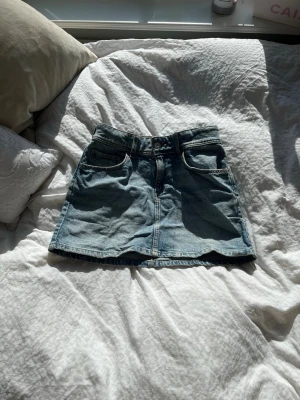  jeanskjol  - Säljer en klassisk blå jeanskjol med normal passform och två framfickor. Kjolen har en kort längd och stängs med knapp och dragkedja framtill. Perfekt till sneakers eller boots för en avslappnad look. Storleken är 152 