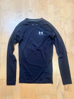 Under armour  träningströja - Svart långärmad träningströja från Under Armour med vit logga på bröstet. Tillverkad i ett stretchigt och fukttransporterande material som passar perfekt för träning och sportaktiviteter. Tight passform och rund halsringning. Storlek S/M