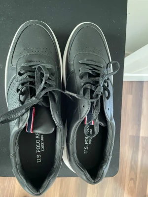 Svarta sneakers från U.S. Polo Assn. - Svarta sneakers från U.S. Polo Assn. med vita sulor och diskreta röda och vita detaljer på plösen. Skorna har klassisk låg modell, perforerade detaljer på tån och ovansidan samt snörning. Tillverkade i syntetmaterial med bekväm innersula.