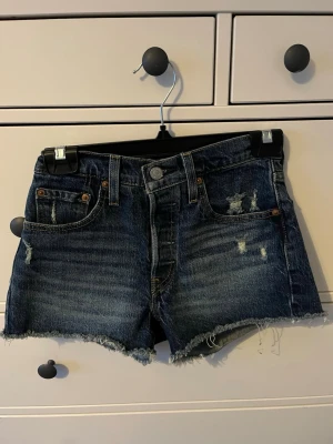 Levi's blå jeansshorts  - Säljer ett par klassiska blå jeansshorts från Levi's med rå, fransig kant och slitna detaljer framtill. Shortsen har fem fickor, knappgylf och en mörk tvätt. Storlek W24 som motsvarar storlek 32.