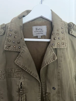 Grön utilityjacka med nitar  - Snygg beige utilityjacka  med coola nitar på kragen och axelklaffar. Jackan har flera fickor, knapp- och dragkedjedetaljer samt en avslappnad passform. Perfekt för dig som vill ha en edgy och trendig look.