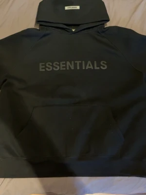 Svart Essentials hoodie med tryck - Svart hoodie från Fear of God Essentials med stort ESSENTIALS-tryck på bröstet och liten logga på huvan. Klassisk känguruficka framtill och ribbade muddar. Perfekt för en clean och stilren streetwear-look.