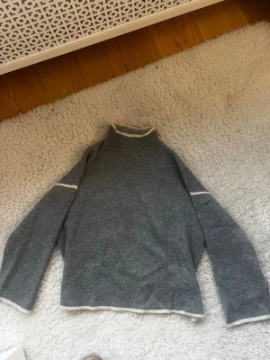 Grå stickad tröja med vita detaljer - Mysig grå stickad tröja med vita kanter vid hals, ärmslut och nederkant. Tröjan har en rak passform, lång ärm och hög hals. Perfekt för lager på lager och enkel att matcha med jeans eller kjol.