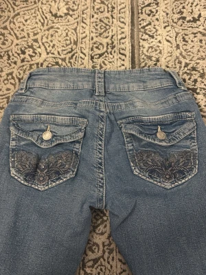 Blå lågmidjade jeans med broderade fickor - Säljer ett par ljusblå jeans med slitningar framtill och snygga broderade detaljer på bakfickorna. I storlek xs från Gina Young 