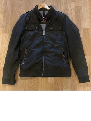 mörkgrön jacka från massimo dutti - Stilren svart vindjacka med hög krage och dragkedja framtill. Två bröstfickor med knappar och två snedställda fickor med dragkedja. Jackan har detaljer i brunt vid kragen och är tillverkad i ett lätt, vindtätt material.