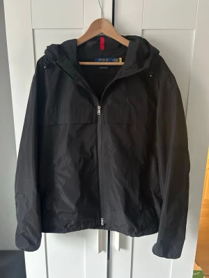Svart vindjacka från Polo Ralph Lauren - Snygg svart vindjacka från Polo Ralph Lauren med huva och dragkedja framtill. Jackan har minimalistisk design, diskret logga på bröstet och justerbar nederkant. Tillverkad i lätt och tåligt material som passar perfekt för blåsiga dagar. Ny pris 3595kr mitt pris 1600kr.                  Pris kan diskuteras. 