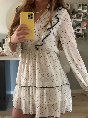 Vit prickig volangklänning Zara - Supersöt kort vit klänning från Zara med svarta prickar, volangdetaljer längs ringningen och kjolen samt v-ringad hals. Klänningen har långa ärmar med lätt puff och tunna, luftiga lager. 