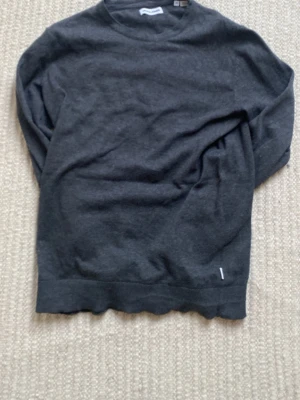 Mörkgrå stickad tröja Jack and Jones - Mörkgrå stickad tröja från Jack and Jones enkel och stilren design. Tröjan har rund halsringning, långa ärmar och ribbade muddar vid ärmslut och nederkant. Perfekt basplagg i mjukt material, troligtvis bomull eller ullblandning.