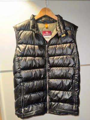 Svart dunväst från Parajumpers - Snygg svart dunväst från Parajumpers med hög krage och dragkedja framtill. Västen har två sidofickor med dragkedja och quiltad design. Insidan är ljusgrå och materialet är glansigt och tåligt. Perfekt för lager-på-lager och streetwear.