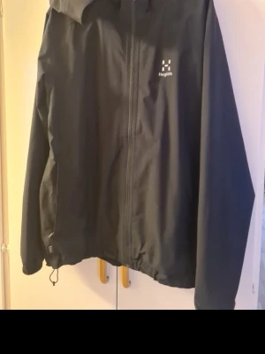 Svart vindjacka från Haglöfs - Ny Snygg svart vindjacka från Haglöfs med huva och dragkedja framtill. Jackan har två sidofickor med dragkedja och justerbar nederkant. Perfekt för blåsiga dagar och enkel att matcha med olika outfits.