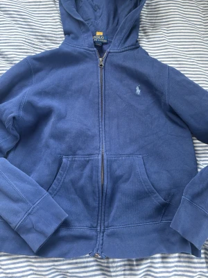 Blå hoodie från Polo Ralph Lauren M (150, 10-12år) - Snygg blå hoodie från Polo Ralph Lauren med dragkedja framtill och klassisk logga broderad på bröstet. Hoodien har huva, två stora fickor fram och ribbade muddar vid ärmar och nederkant. Tillverkad i mjukt bomullsmaterial för skön känsla. Junior storlek M (10-12 år)